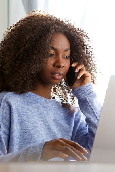 young black woman listens on phone while looking at laptop