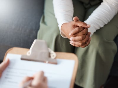 A woman's folded hands rest across from her health care provider who holds a clipboard during their consultation.